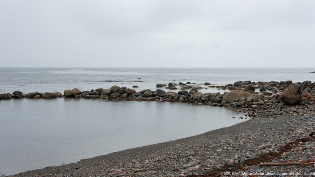 Small rocky cove with a curved dark beach and low boulder breakwater extending across gray coastal water.