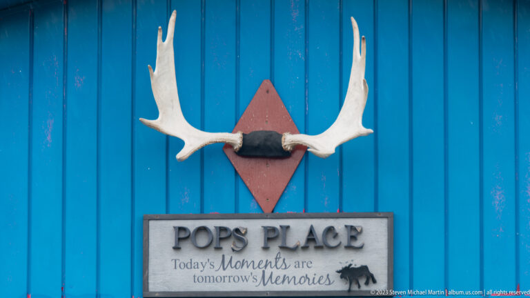 Pale antlers mounted on bright blue siding above a rustic “Pop’s Place” sign with a sentimental motto.