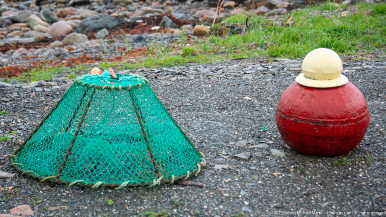 Conical green mesh fishing trap beside a large red buoy with a pale yellow top on gravel.