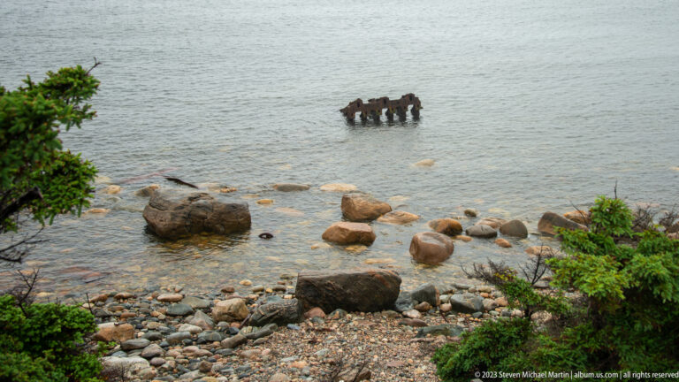 Old wooden pilings stand in shallow gray water beyond a cobble beach with scattered boulders and spruce branches.