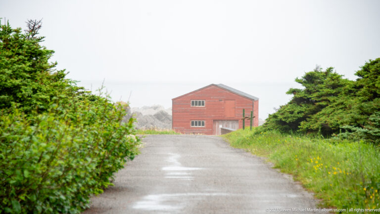 Red gabled utility building at the end of a wet gravel road, framed by shrubs and spruce near the coast.