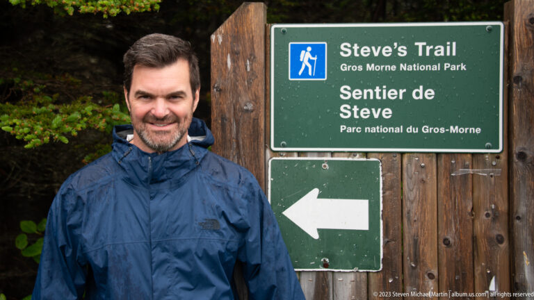 Smiling man in a blue rain jacket beside the bilingual “Steve’s Trail” sign and left-pointing arrow.