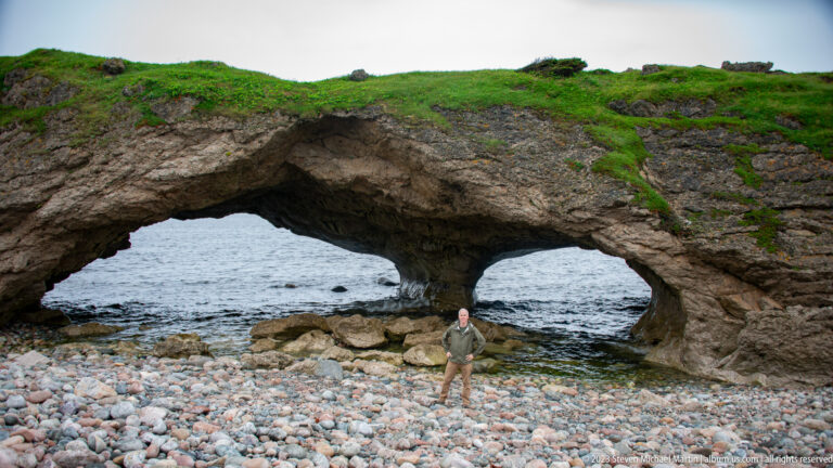 Two-opening coastal rock arch above a cobble beach, with a person standing beneath the central span for scale.