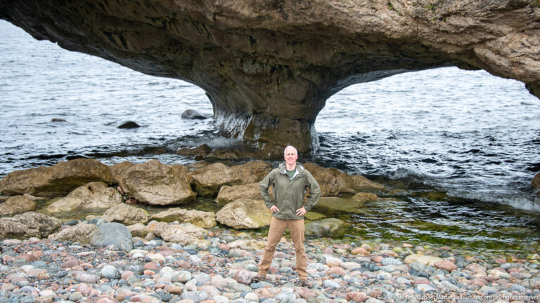 Person standing on a cobble beach beneath a double coastal rock arch with waves visible through the openings.