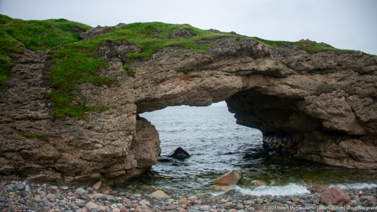 Large natural rock opening framing the gray sea, with green turf above and waves entering over a cobble shore.