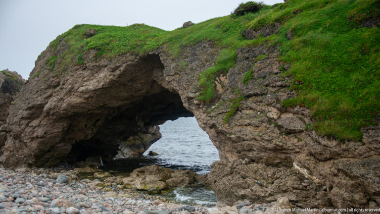 Oblique view of a natural sea arch beneath a grassy rock headland with water flowing through at beach level.