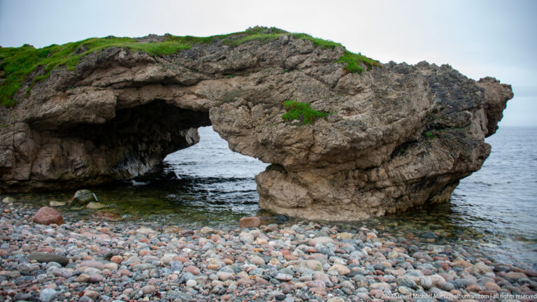 Low rugged sea arch beside a broad cobble beach, with gray ocean visible through the opening and grass above.