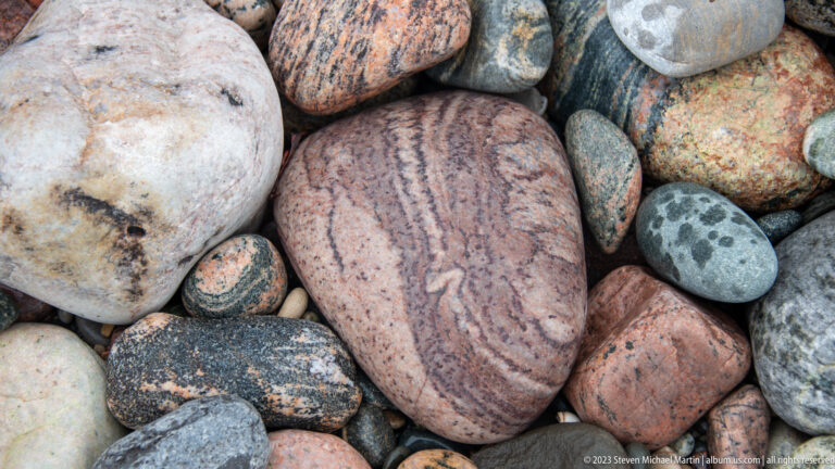 Large pink rounded cobble with dark wavy bands surrounded by smaller multicolored stones on a beach.