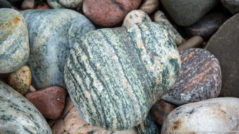 Large green-gray rounded cobble with dense pale bands among darker multicolored beach stones.