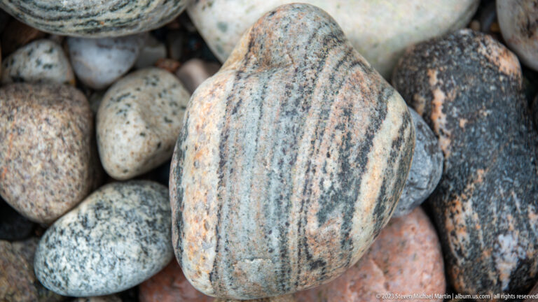 Rounded gray cobble with curved dark, pale, and salmon-pink bands surrounded by other beach stones.