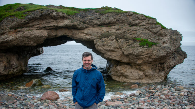 Man in a blue rain jacket standing on a cobble beach in front of a broad natural sea arch.