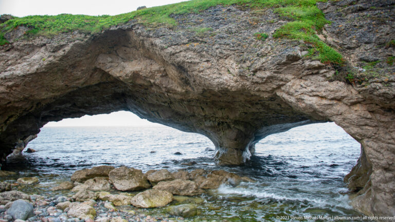 Double coastal rock arch with a narrow central pier, waves beneath the openings, and boulders across the foreground.
