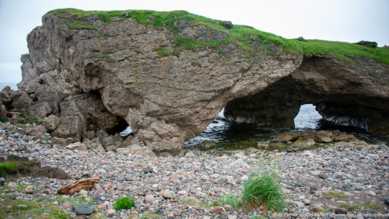 Fractured coastal rock arch seen from the side above a cobble beach, with seawater visible beneath the right opening.