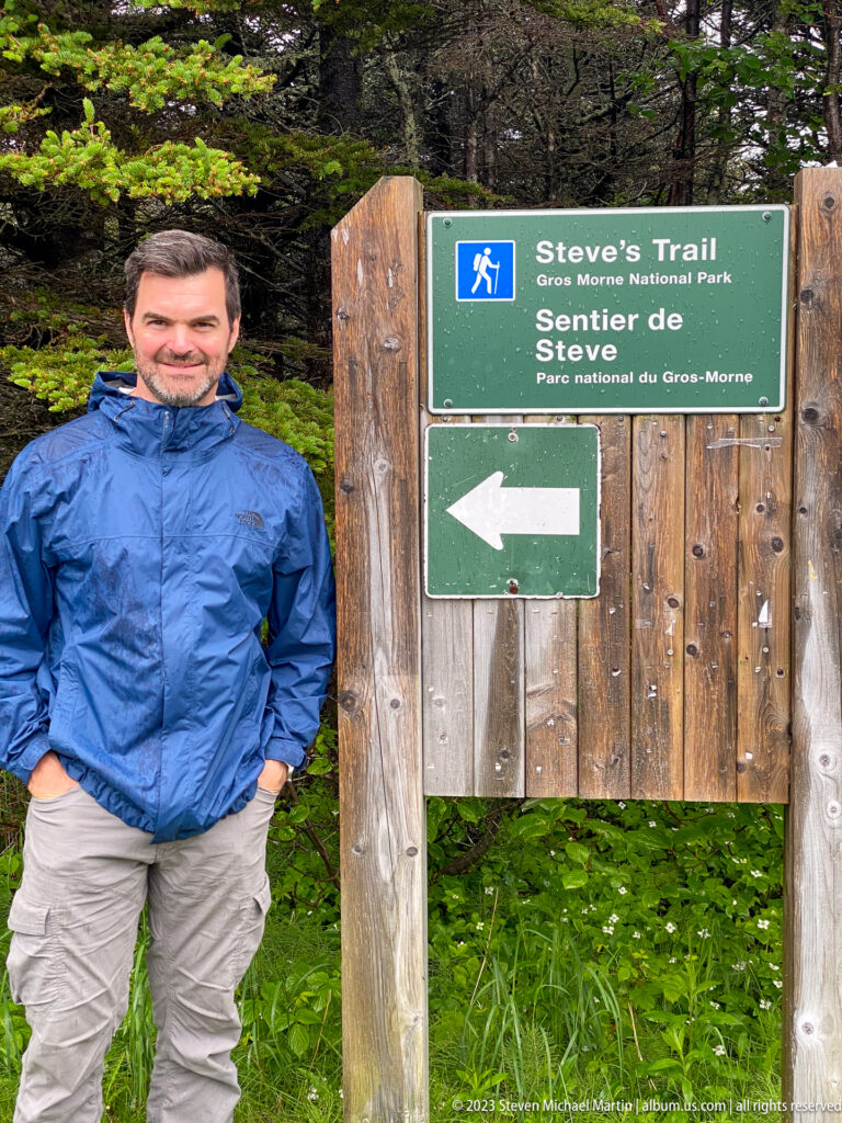 Man in a blue rain jacket beside the bilingual Steve’s Trail sign and left-pointing arrow in dense forest.
