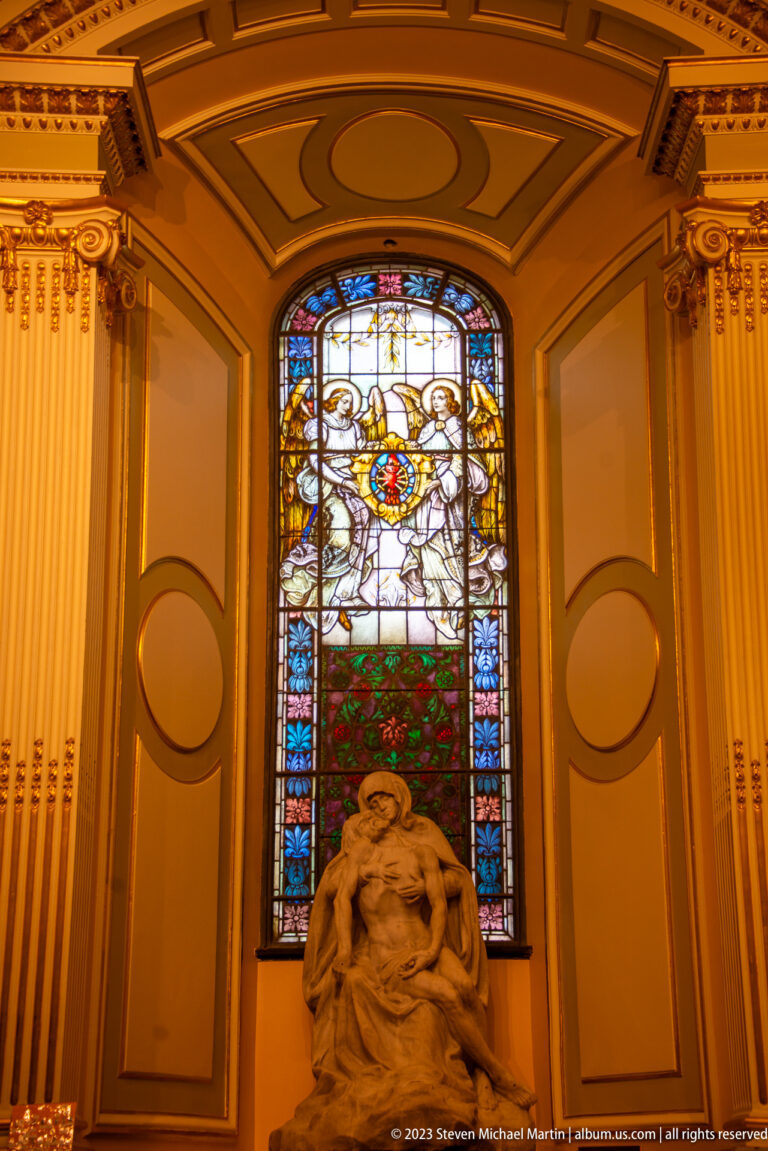 Pale Pietà-like sculpture of Mary supporting Christ beneath an ornamental stained-glass window with a central emblem.