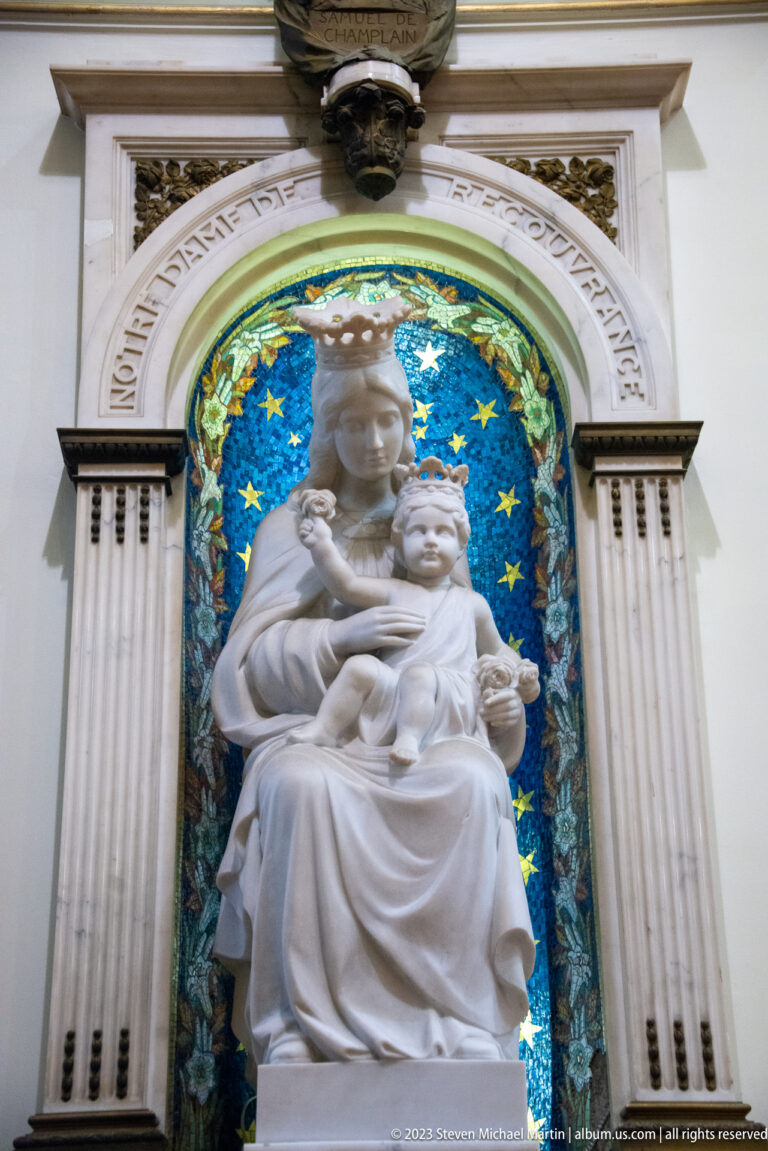 White seated Virgin and Child statue in a classical niche backed by a deep blue mosaic filled with gold stars.