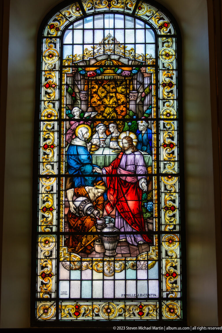 Stained-glass window with three principal biblical figures in a domestic interior, framed by an ornate gold-and-floral border.