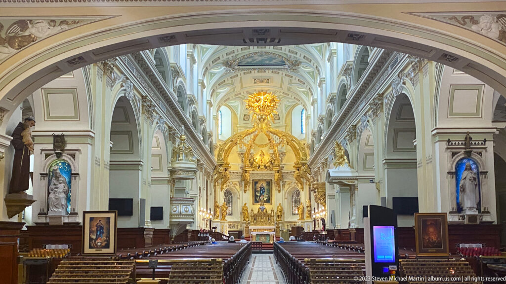 Centered nave view beneath an arch, showing pews, side niches, pale vaulting, and the gilded sanctuary at the far end.