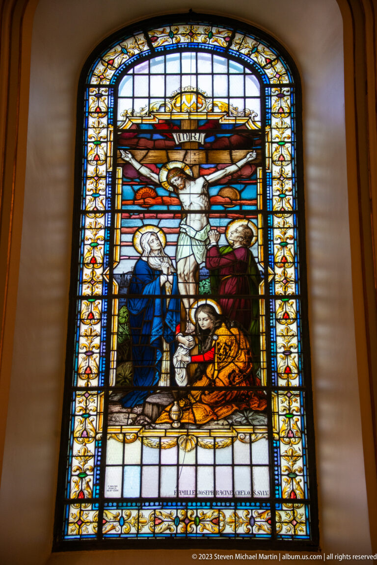 Tall stained-glass Crucifixion scene with Christ on the cross and mourners gathered below against a red-orange sky.