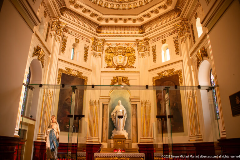 Small cream-and-gold chapel with a central Marian statue, large religious paintings, classical ornament, and a glass barrier in front.