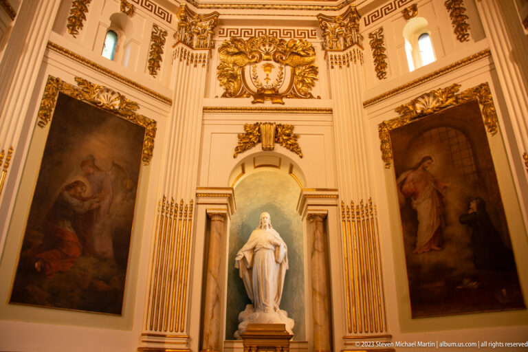White Virgin statue in a central niche between two large religious paintings, surrounded by cream-and-gold architectural ornament.