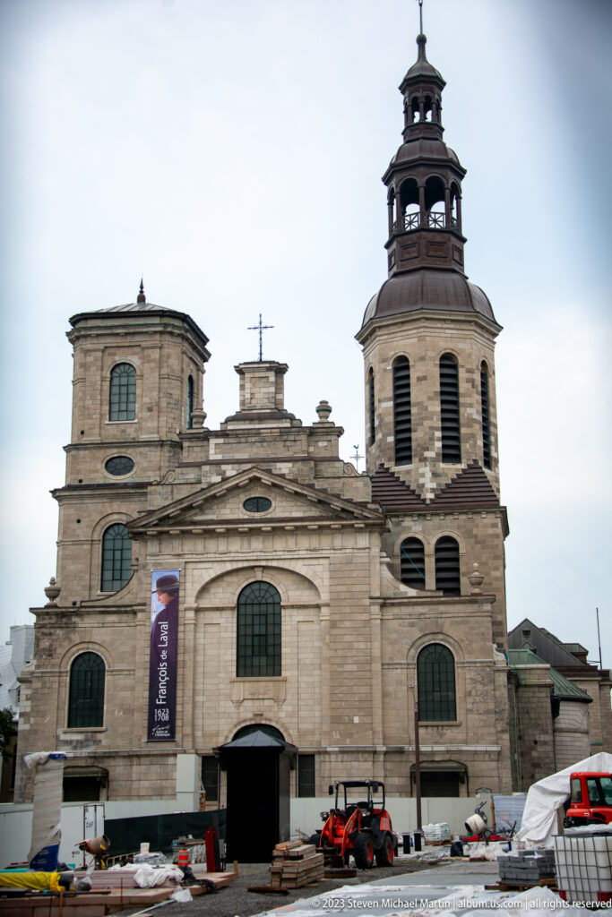 Front façade of Notre-Dame de Québec with pale stonework, arched windows, tall bell tower, and construction activity at street level.