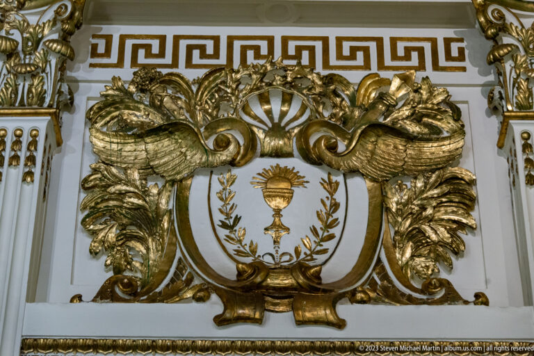 Gilded relief with a chalice and radiant host inside a wreath, surrounded by scrolling foliage beneath a Greek-key border.