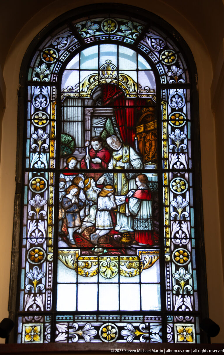 Arched stained-glass window showing robed clergy beneath a red canopy with a gathered crowd inside an architectural setting.