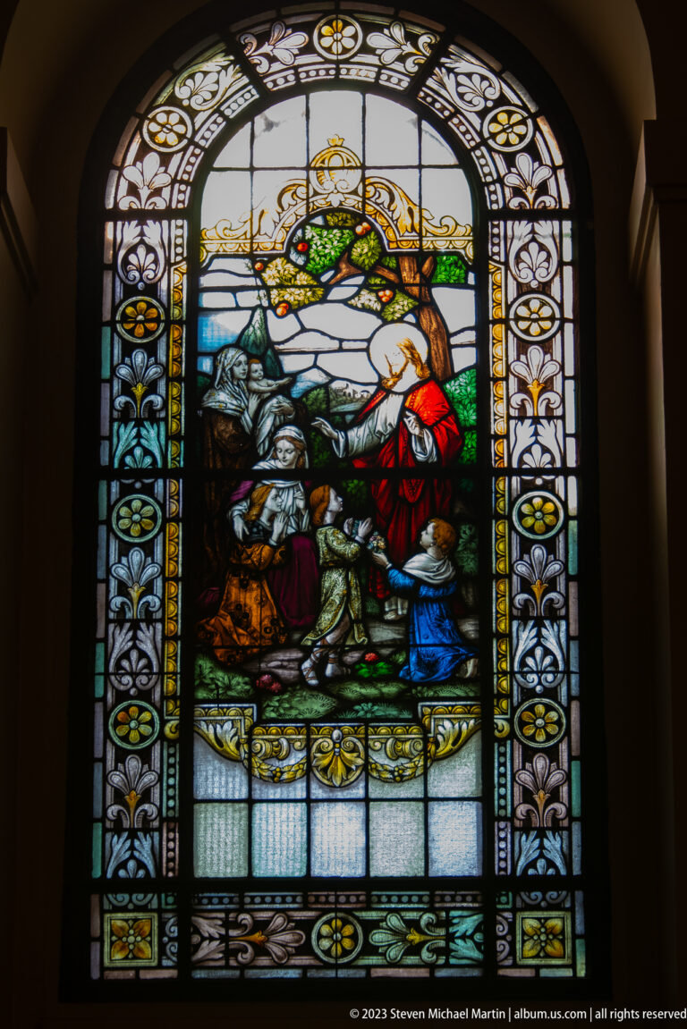 Arched stained-glass religious scene with a red-robed standing figure and kneeling companions in a rocky mountain landscape.