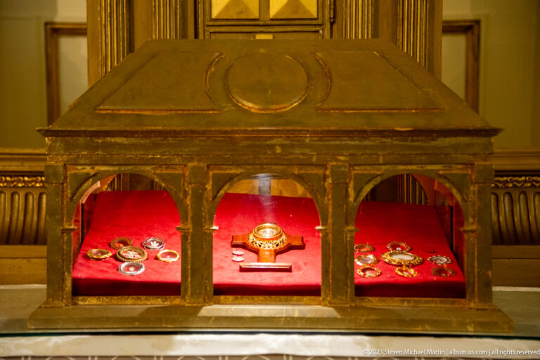 Dark three-bay glass display case holding small devotional or relic objects on bright red fabric.