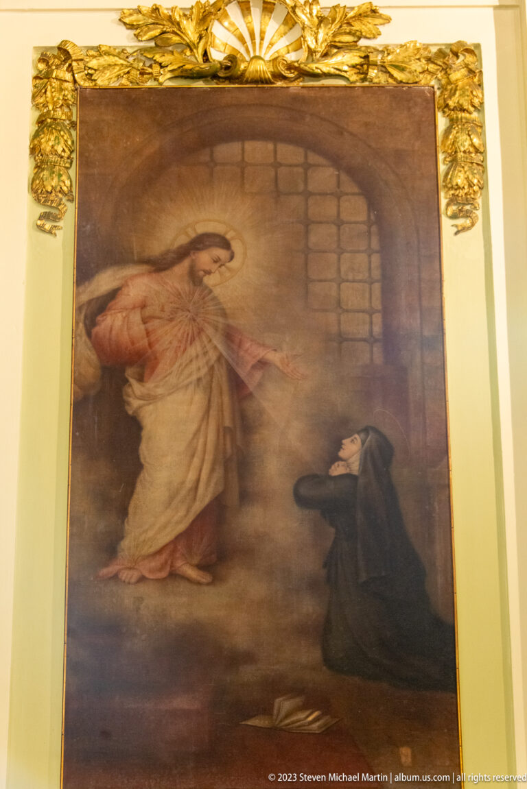 Religious painting with a robed male sacred figure gesturing toward a kneeling woman in a dark habit beside masonry walls.
