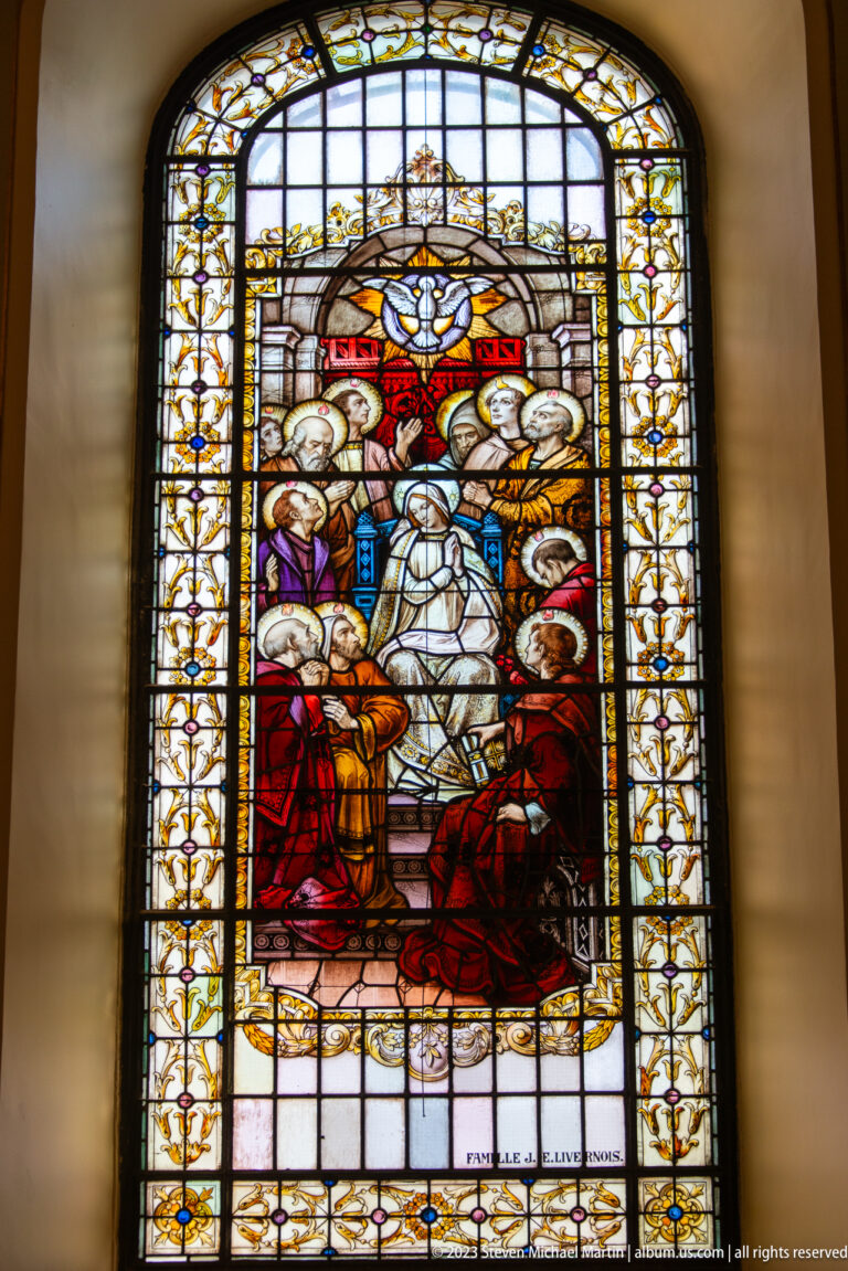 Stained-glass window with a white-robed cleric surrounded by clergy in red and purple, beneath a radiant dove-like symbol.