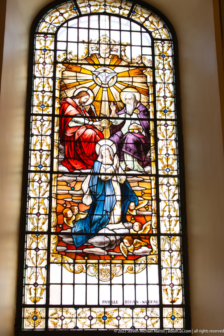 Stained-glass scene with a radiant white dove above a seated woman in blue and gathered figures, recalling Pentecost iconography.