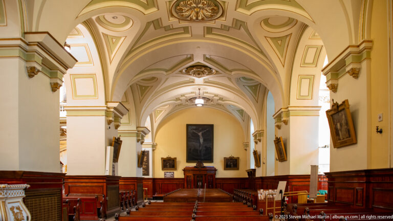 Long side aisle with wooden pews, cream vaulted arches, framed religious paintings, and dark wood wainscoting.