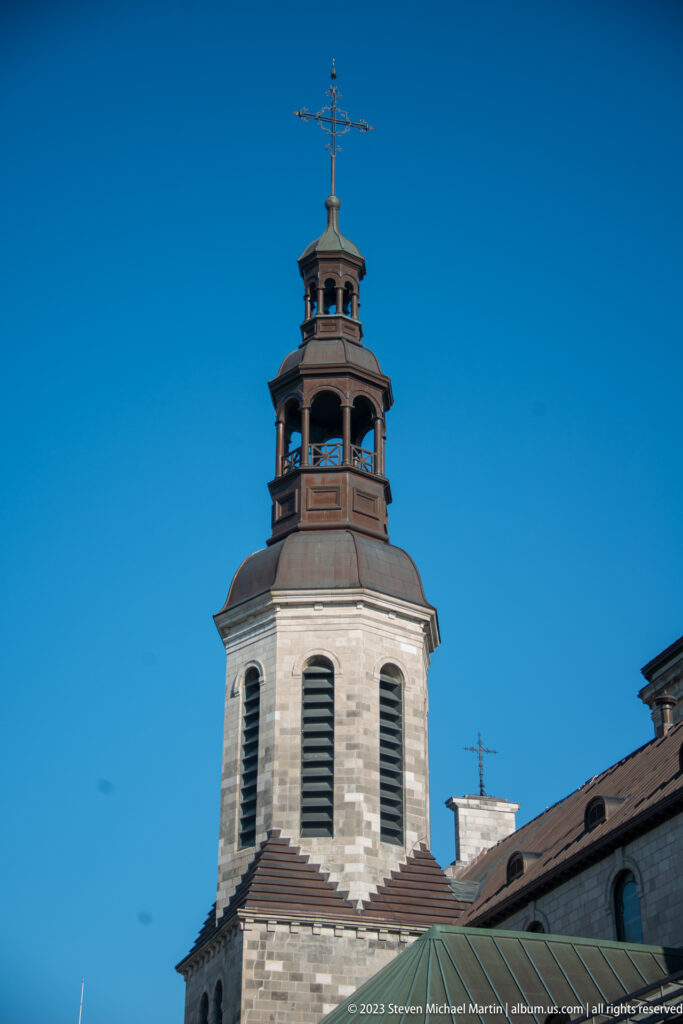 Close view of Notre-Dame de Québec's stone bell tower with louvered openings, open belfry, and cross against blue sky.