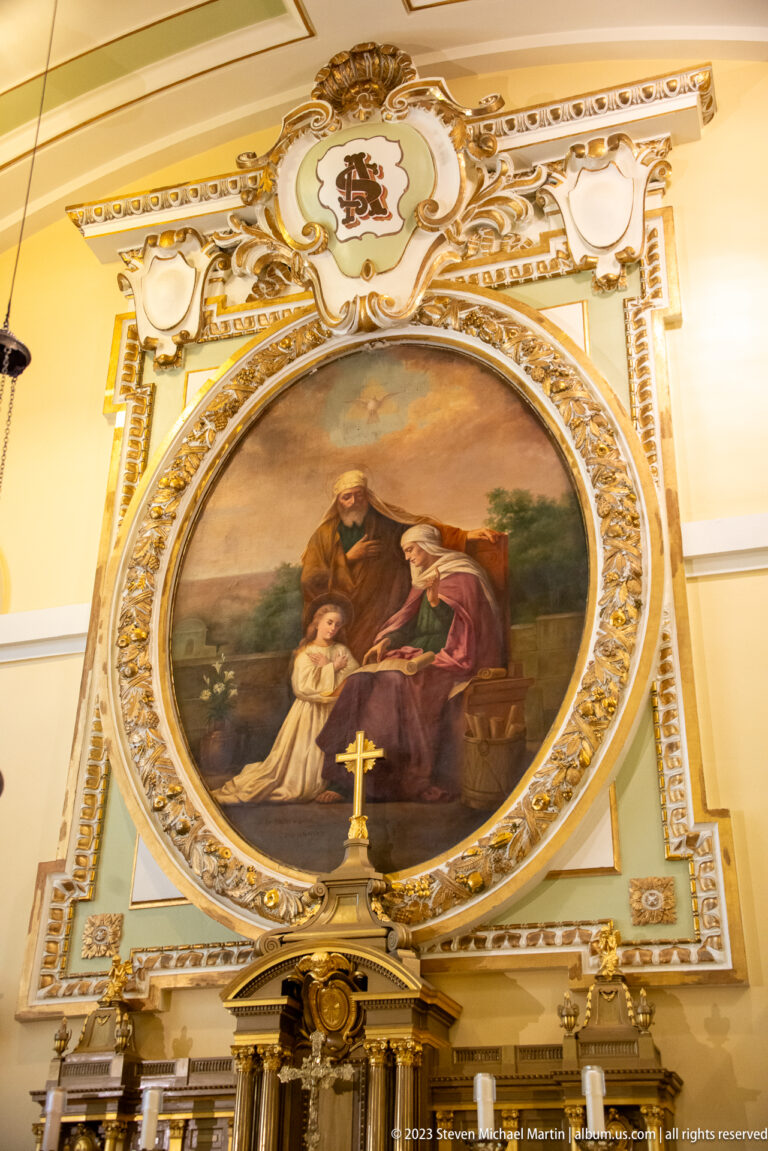Oval religious painting with several figures in an ornate cream-and-gold frame above the top of a side altar.