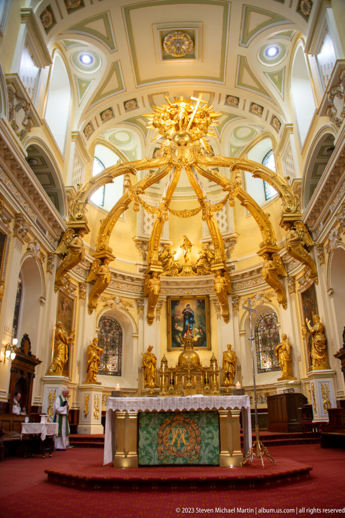 Wide basilica sanctuary with high altar, gilded canopy, pale vaulted ceiling, red carpet, and sculptural decoration.