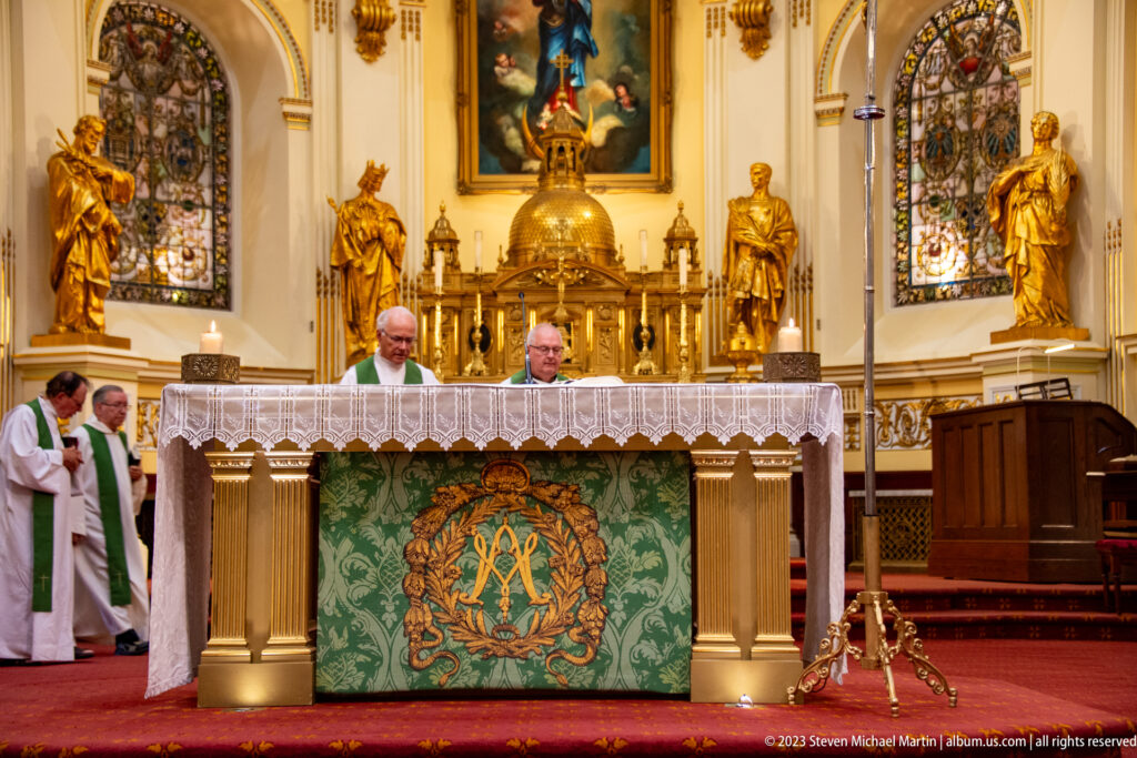 Clergy behind a lace-covered altar with green embroidered frontal, gilded statues, tabernacle, stained glass, and painting.