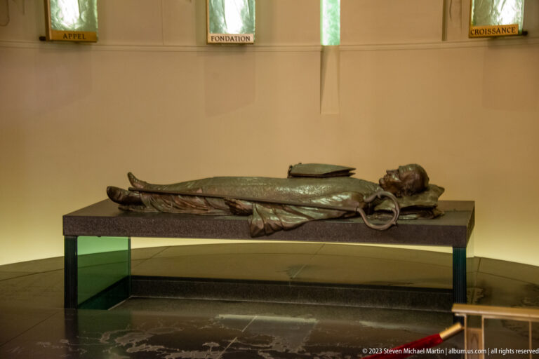 Life-size dark recumbent figure on a low platform beneath narrow windows in a pale church interior.