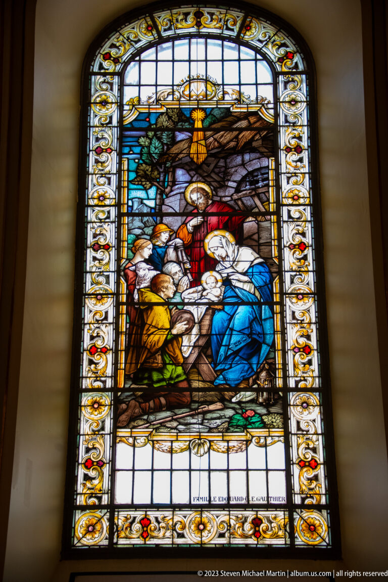 Tall stained-glass window with kneeling and standing figures around a blue-robed woman, framed by floral borders.