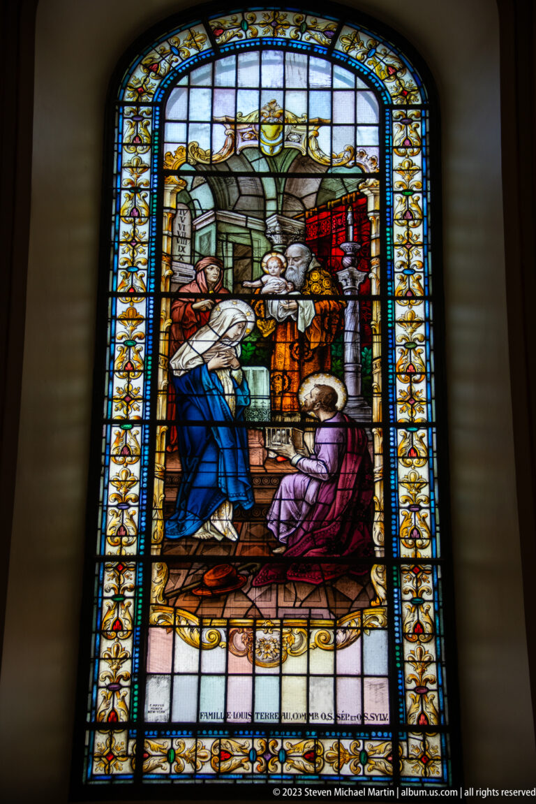 Stained-glass scene with a kneeling woman facing a group of figures amid architectural details and floral borders.