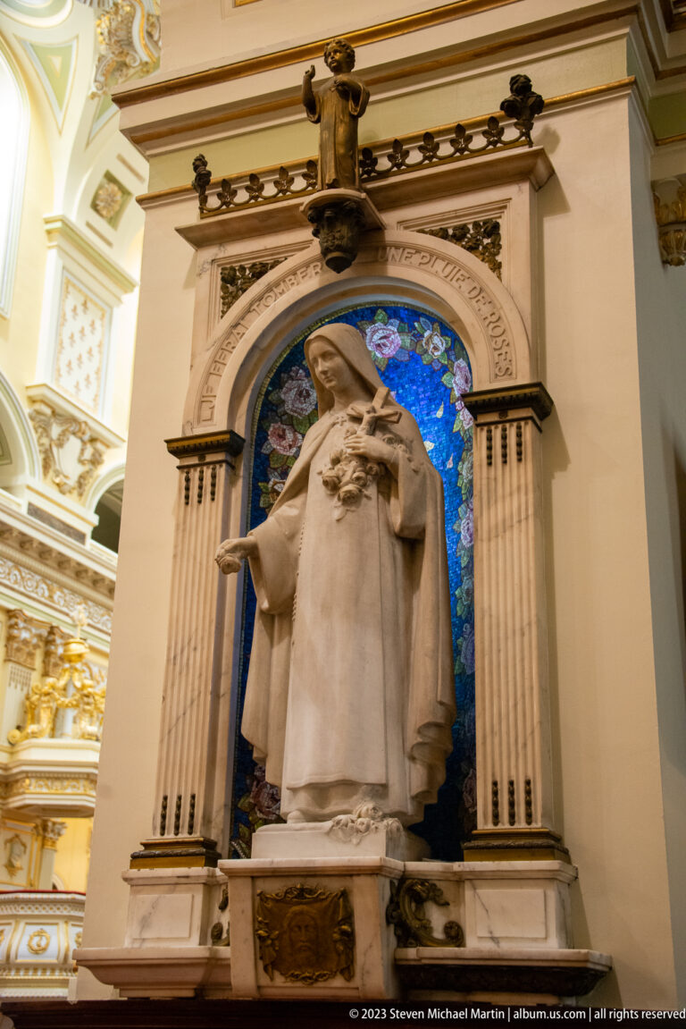 White stone statue of a veiled female saint in a carved niche backed by vivid blue mosaic.