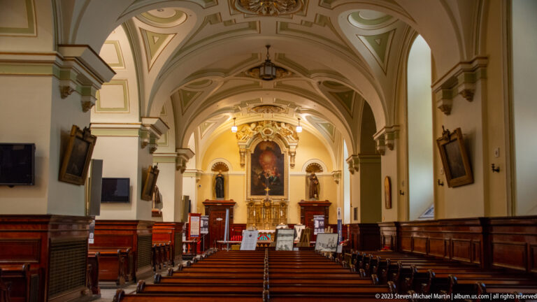 Long church interior with wooden pews, pale arches and vaults, devotional pictures, and an altar at the far end.