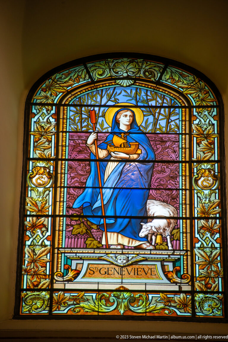 Stained-glass Saint Geneviève holding a staff with a white sheep beside her, identified by an inscription below.