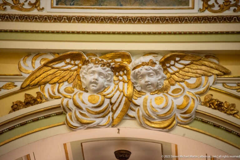 Pair of white cherub heads with gilded wings and scrollwork mounted above a cream interior arch.