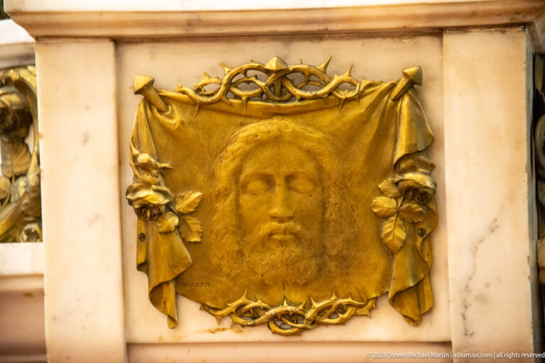 Gilded relief of Christ’s face on a draped cloth, encircled by thorn-like ornament in a pale stone panel.