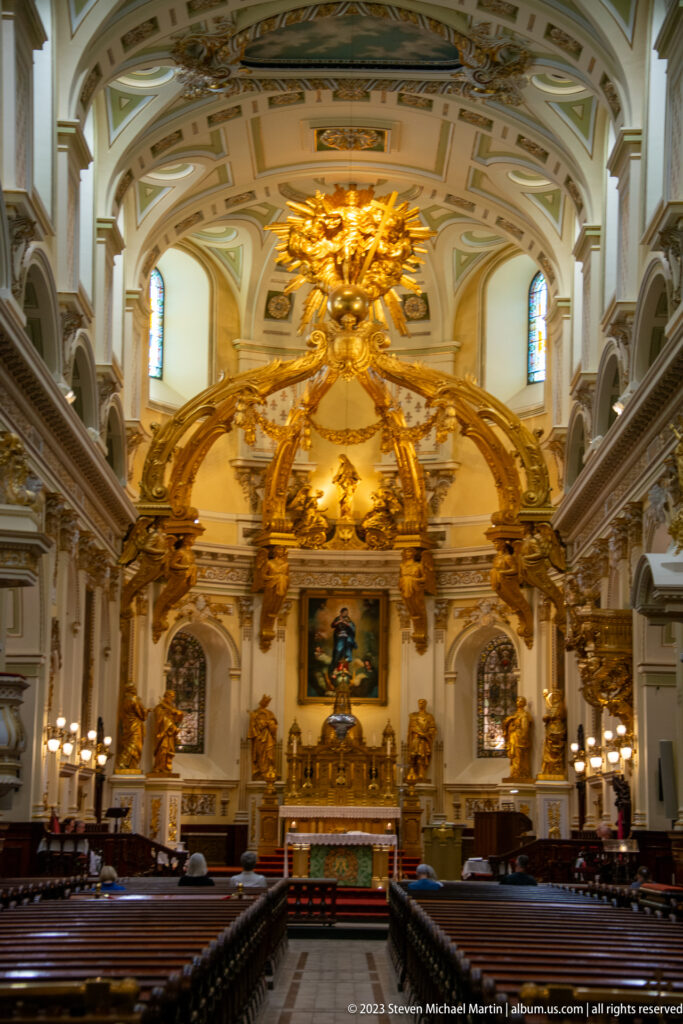 Wide nave view toward a monumental gilded sanctuary canopy and high altar, framed by arches and stained-glass windows.