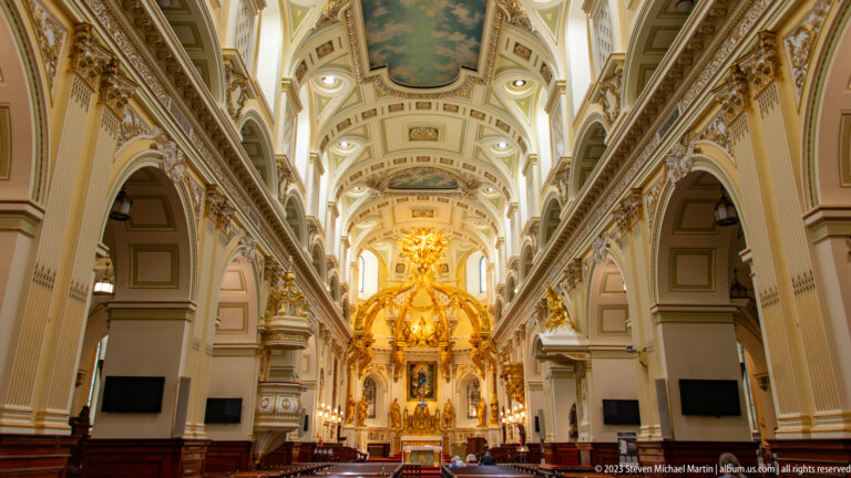 Closer view along Notre-Dame de Québec's nave toward the gilded altar, framed by cream arcades, painted vaulting, and dark wood furnishings.