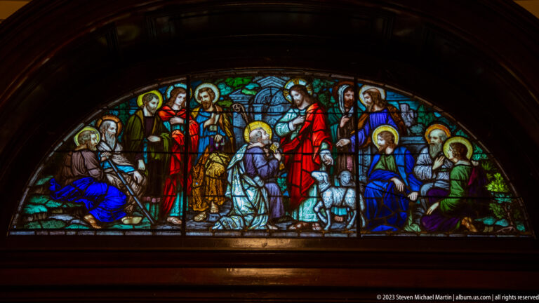 Semicircular stained-glass window with numerous haloed biblical figures in red, blue, green, and gold, set within dark woodwork.