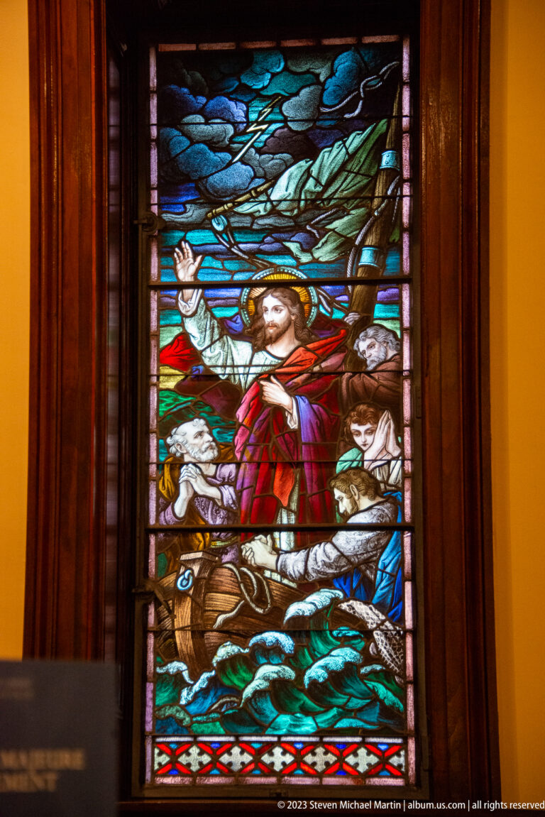 Vertical stained-glass panel of Christ in red amid turbulent blue-green water, with other figures and a vessel-like form below.